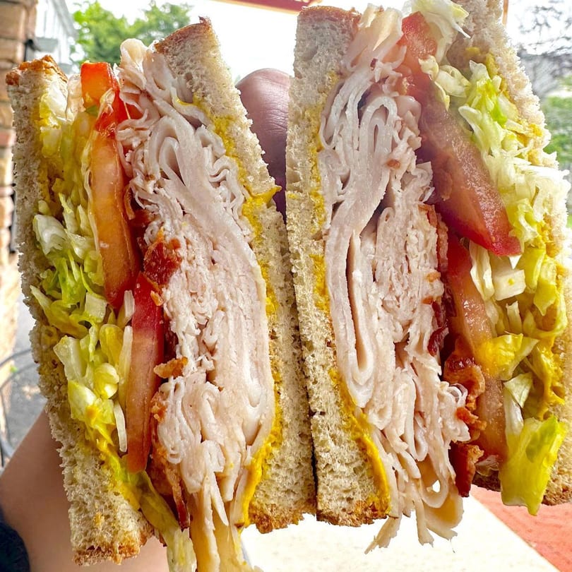 A large turkey sandwich from Sandwich Club, cut in half, revealing layers of meat, lettuce, tomato, and mustard.