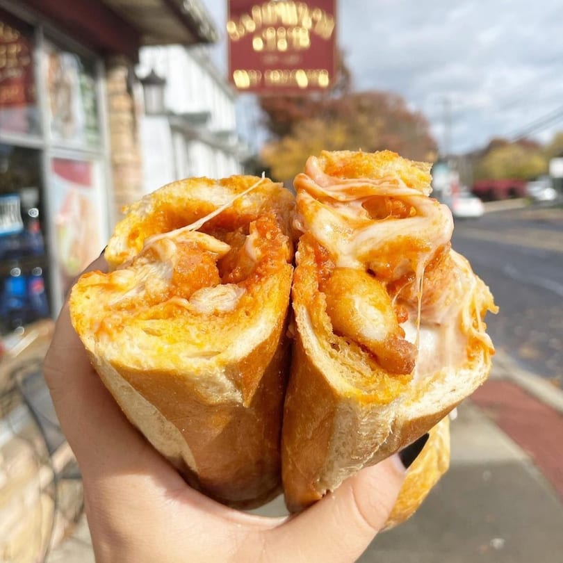 A hand holds a buffalo chicken sandwich, cut in half, with melted cheese and sauce, outdoors.